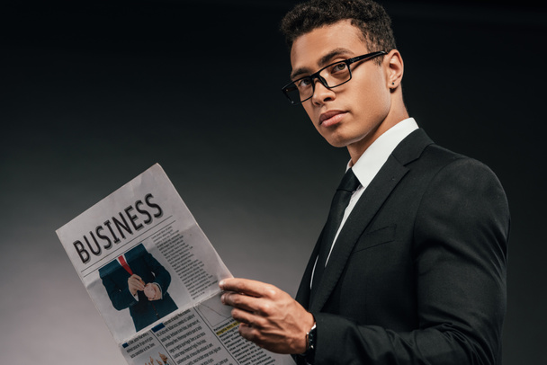 vista lateral del hombre de negocios afroamericano en gafas y traje leyendo periódico de negocios sobre fondo oscuro
 - Foto, Imagen