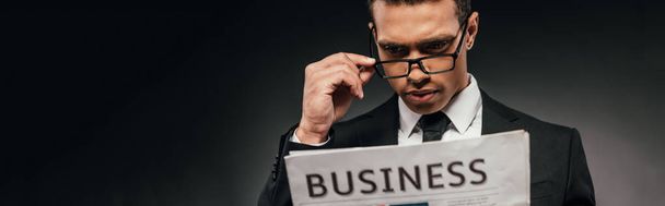 handsome african american businessman in glasses and suit reading business newspaper on dark background, panoramic shot - Фото, изображение
