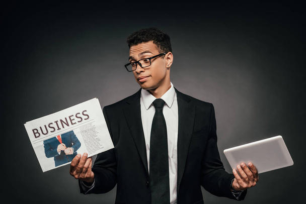 confused african american businessman in glasses and suit holding business newspaper and digital tablet on dark background - Photo, image