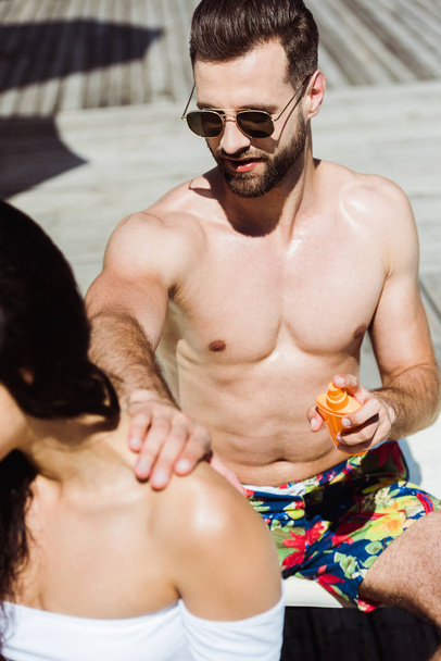selektiver Fokus eines gutaussehenden Mannes mit Sonnenbrille, der junge Frau berührt und Flasche mit Sonnencreme in der Hand hält  - Foto, Bild