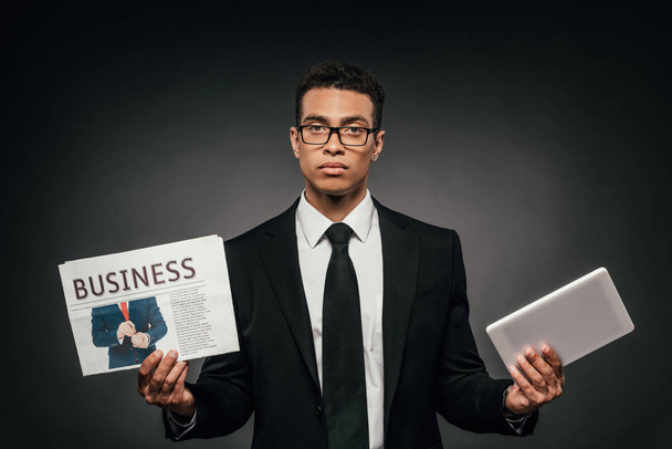 empresário afro-americano em óculos e terno segurando jornal de negócios e tablet digital em fundo escuro
 - Foto, Imagem