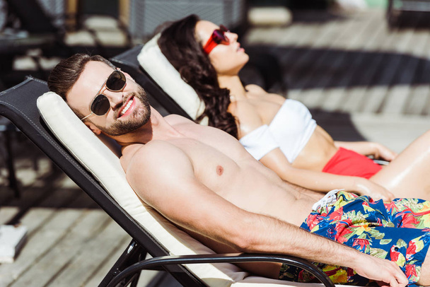 selective focus of happy muscular man in sunglasses lying on deck chair near woman  - Photo, Image