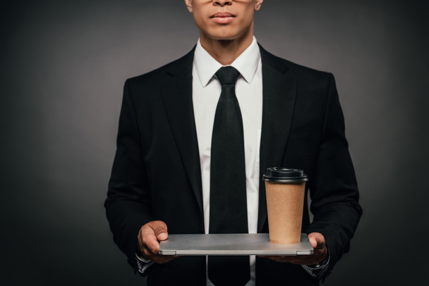 vista recortada del hombre de negocios afroamericano sosteniendo portátil y taza de papel sobre fondo oscuro
 - Foto, Imagen