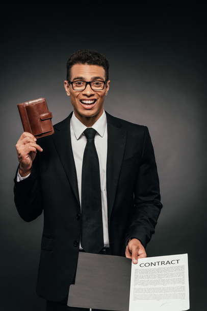 vzrušený afroamerický podnikatel zobrazující kontrakt a držení kožených peněženky na tmavém pozadí - Fotografie, Obrázek