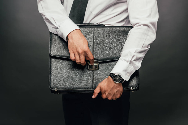 cropped view of african american businessman holding leather briefcase on dark background - Photo, Image