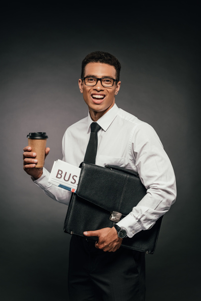 happy african american businessman holding newspaper, coffee to go and leather briefcase on dark background - Fotoğraf, Görsel