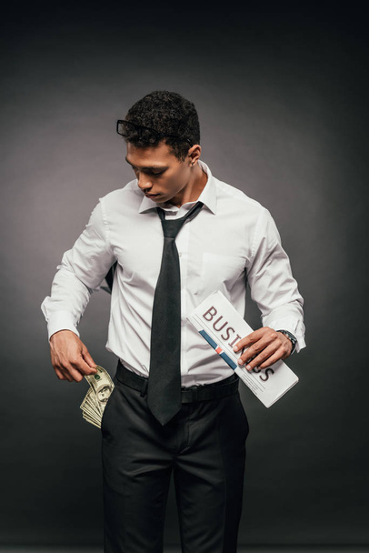 Αφρικανός Αμερικανός επιχειρηματίας με μετρητά στην τσέπη που κατέχουν επιχειρηματική εφημερίδα σε σκοτεινό φόντο - Φωτογραφία, εικόνα