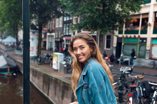 Beautiful face of young smiling woman. White teeth, big lips. Curly hairstyle, modern makeup. Blue jeans jacket. Standing on the bridge, enjoys the river flow. Bicycles on the background. - Photo, Image