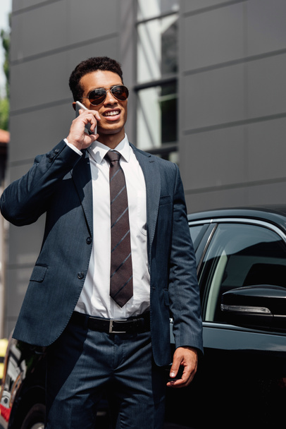 african american businessman in suit and sunglasses talking on smartphone at sunny day near car - Fotoğraf, Görsel
