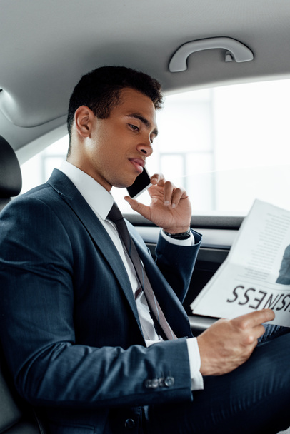 african american businessman reading business newspaper and talking on smartphone in car - Zdjęcie, obraz