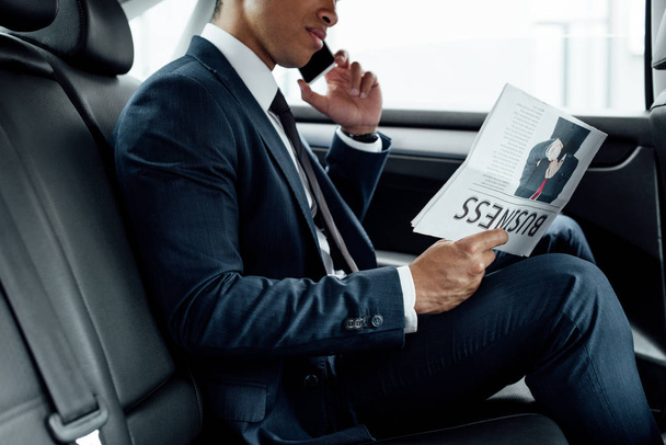partial view of african american businessman reading business newspaper and talking on smartphone in car - Foto, Imagen