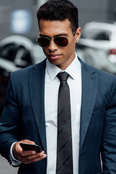 handsome african american businessman in suit and sunglasses using smartphone - Фото, изображение