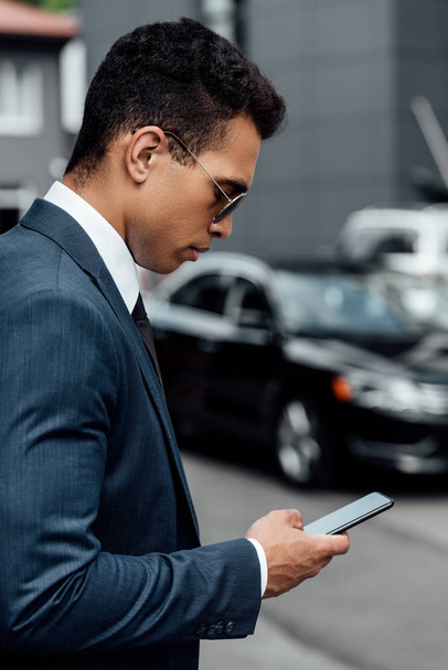 side view of handsome and confident african american businessman in suit and sunglasses using smartphone - Photo, Image