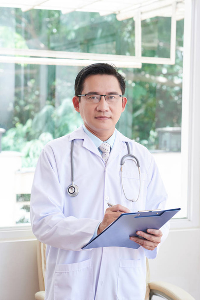 Portrait of Asian male doctor in white coat holding clipboard and writing prescription in hospital - Foto, Bild