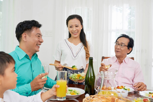 Jovem mulher asiática servindo salada fresca para homens adultos no jantar de família enquanto se reúne à mesa
 - Foto, Imagem
