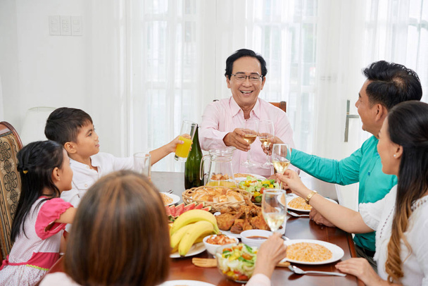 Adultos asiáticos con niños reunidos en la mesa en casa mientras brindan con gafas y celebran
 - Foto, Imagen