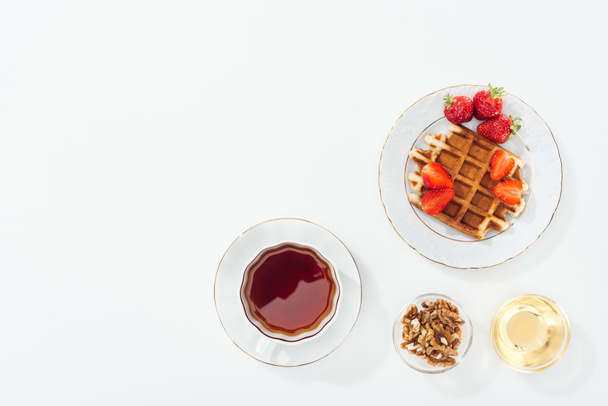 сверху вид на вафли с клубникой на тарелке возле чашки с чаем, миски с медом и орехи на белом
 - Фото, изображение