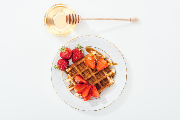 top view of bowl with honey and wooden dipper near plate with waffle and strawberries on white - Valokuva, kuva
