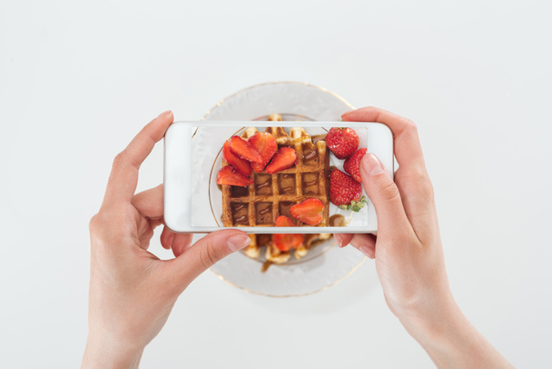 cropped view o woman taking picture of tasty waffle with strawberries on plate isolated on white - Fotó, kép