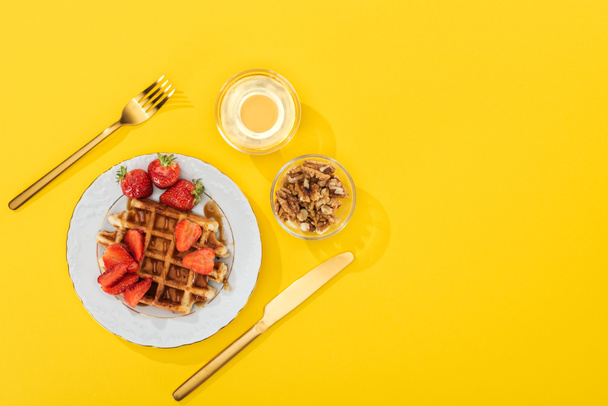 сверху вид на подаваемый завтрак с вафлями, ягодами, медом и орехами возле столовых приборов на желтый
 - Фото, изображение
