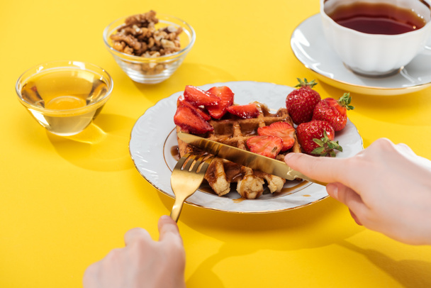 cropped view of woman cutting waffles on plate near bowl and cup with tea on yellow background - Foto, Imagen