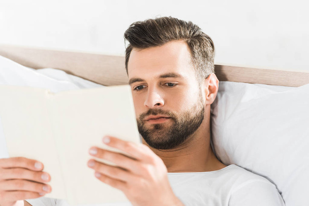 handsome man lying in bed and reading book in morning - Foto, afbeelding