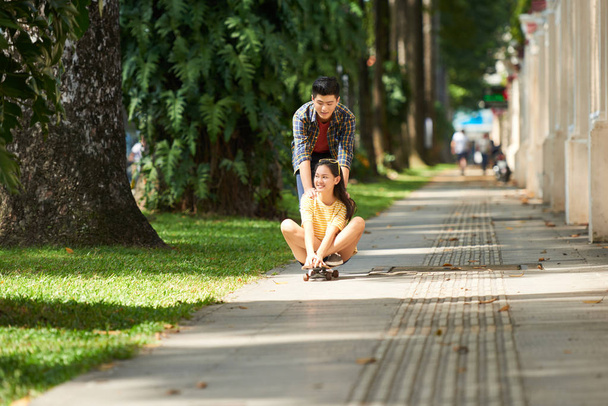 fröhliches junges asiatisches Paar reitet gemeinsam auf Skateboard im Park  - Foto, Bild