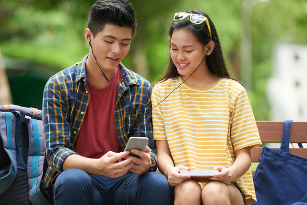 fröhliches vietnamesisches Paar teilt Kopfhörer beim Ansehen von Musikvideos auf dem Smartphone-Bildschirm - Foto, Bild