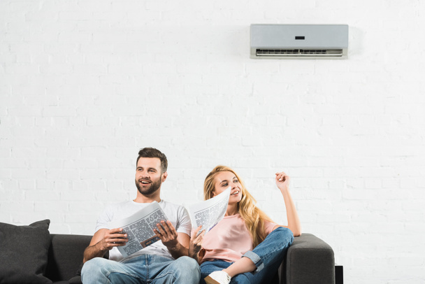 couple on couch with newspapers suffering from heat under air conditioner at home - Photo, Image
