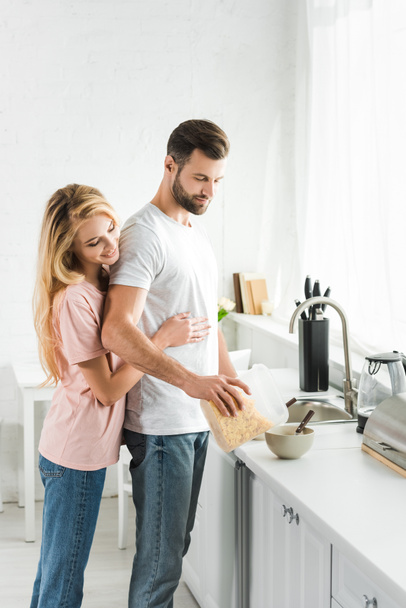 γυναίκα αγκαλιάζει άνθρωπος ρίχνει δημητριακά κατά τη διάρκεια του πρωινού στην κουζίνα - Φωτογραφία, εικόνα