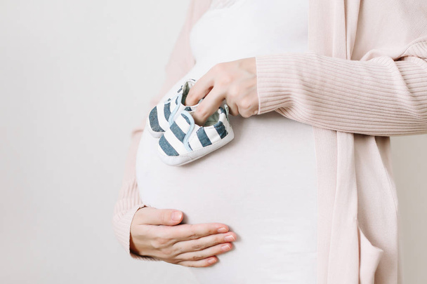 Belly of a pregnant woman in white clothes, mother hands holding shoes for the newborn baby - Photo, image