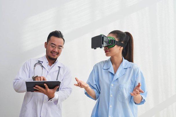 Улыбающийся азиатский врач контролирует приложение виртуальной реальности, которое тестирует его коллега-женщина
 - Фото, изображение