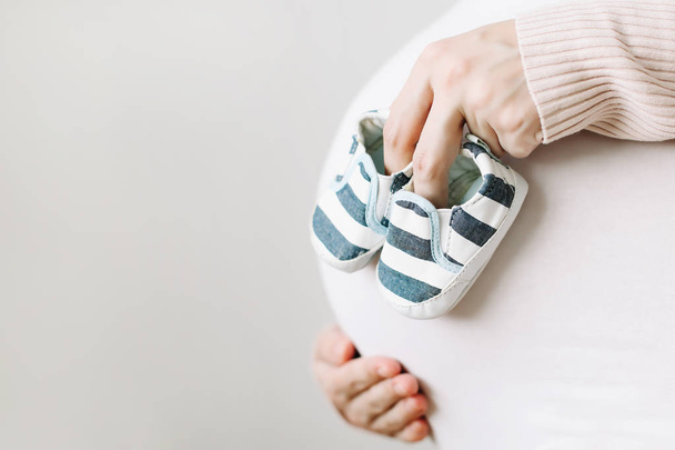 Břicho těhotné ženy v bílých šatech, matky držící boty pro novorozené dítě - Fotografie, Obrázek
