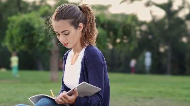 Calm brunette woman sitting on grass and writing something in notebook in park - Video