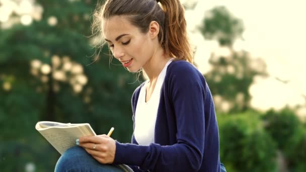 Pleased brunette woman sitting on grass and writing something in notebook in park - Footage, Video