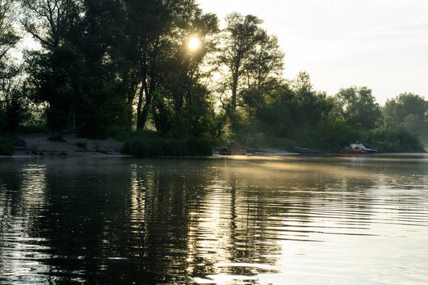 All'alba. Il sole splende tra gli alberi e la luce del sole si riflette nel fiume. Sul fiume c'è una nebbia. Ci sono barche vicino alla riva. Concetto - viaggiare
. - Foto, immagini