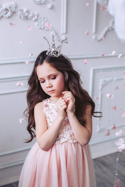 Cute dark-haired girl is embarrassed, posing in a bright interior against a white wall in a beautiful pink dress and a crown on her head. - Photo, Image