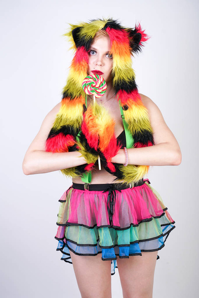 charming plus size young woman in fur hat made of multicolored fibers with cat ears and paws posing in green suspenders, black bra and funny short rainbow skirt on a white background in the Studio - Foto, immagini