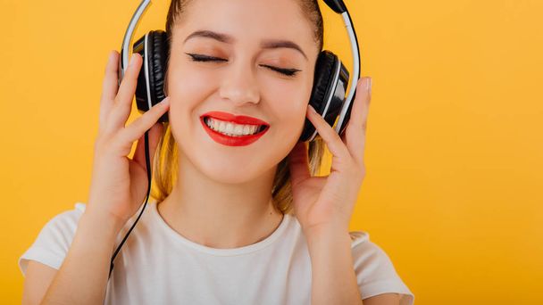 cropped. girl smiling with eyes closed, listening to music headphones on head dressed in a white shirt, positive emotional state  isolated on a yellow background - Fotografie, Obrázek