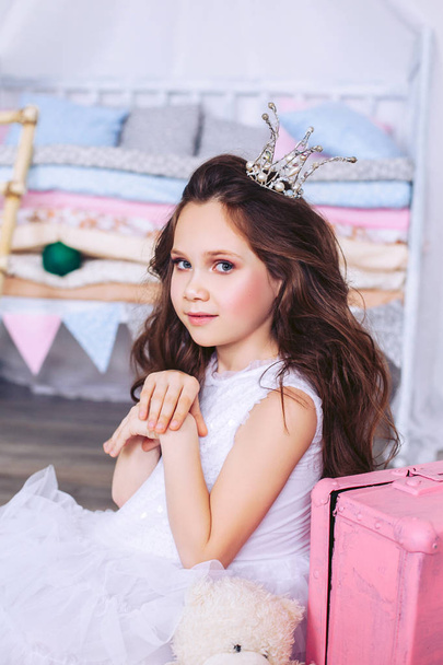 A young brunette with a crown on her head is sitting on the floor next to a pink suitcase, in a white beautiful dress. - Foto, Imagem