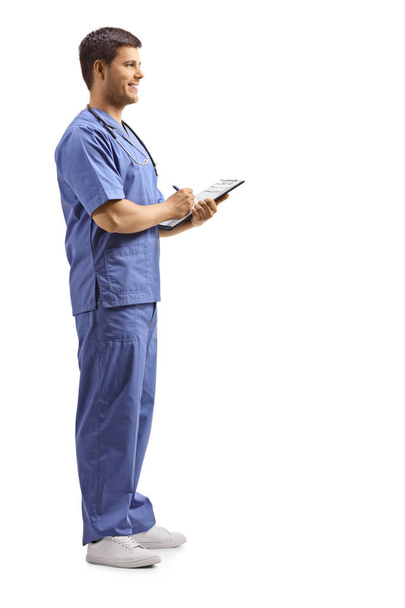 Full length profile shot of a young male health pratctitioner in a blue uniform writing on a clipboard isolated on white background - Foto, Bild