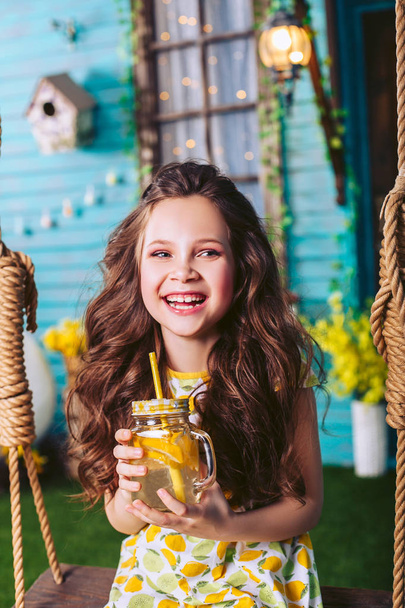 Little dark-haired girl laughs sitting on a swing, holding lemonade in her hands - Valokuva, kuva