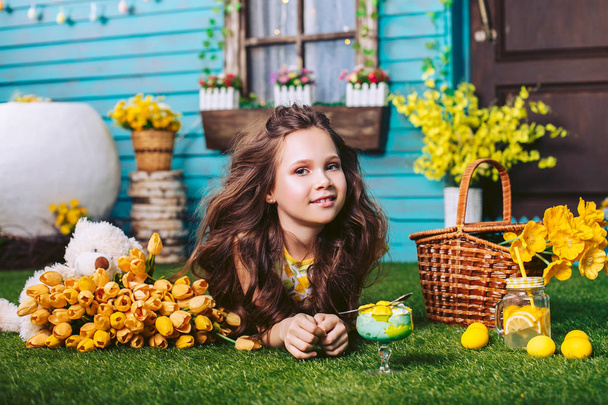 A girl with voluminous curls lies on a green lawn and smiles against the background of a wooden blue house. Celebration of spring and Easter. - 写真・画像