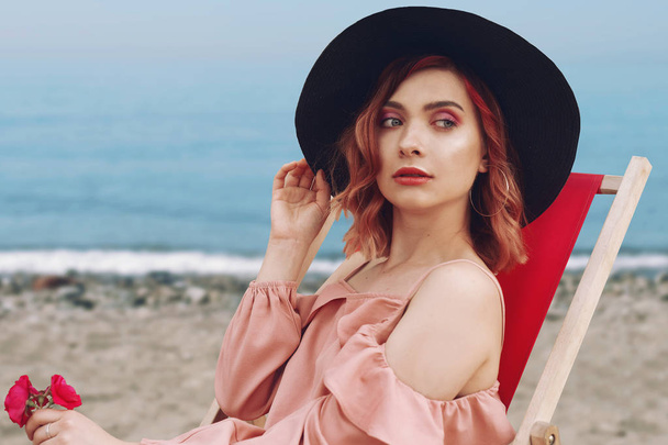 Beautiful girl in a hat with pink hair and beautiful makeup lying on a lounger against the beach and the ocean. Beautiful girl resting on the beach on a lounger on the background of the sea. Fashion. - Fotografie, Obrázek