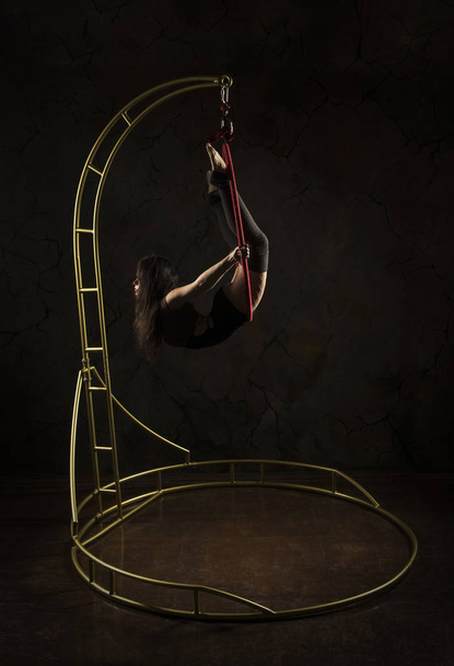 Gorgeous girl, aerial acrobat, in a dark suit performs acrobatic elements on a portable air ring, in the stage light. - Foto, imagen