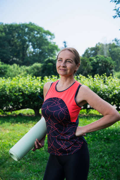 Женщина 50 лет, стоящая в парке с ковриком для йоги
 - Фото, изображение