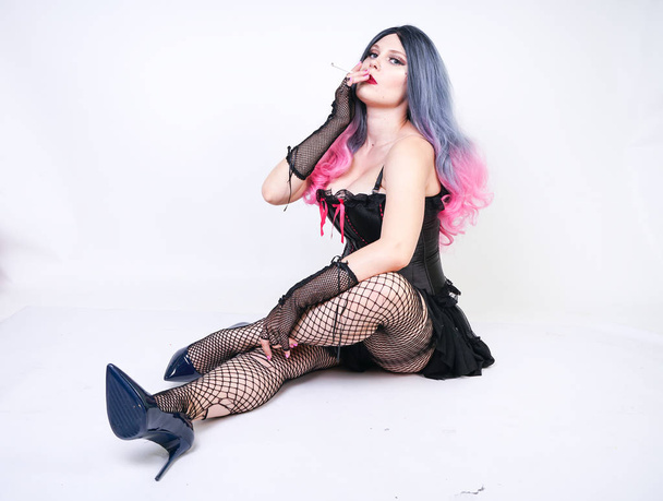 κακό κορίτσι με διάτρηση και μακριά γκρίζα ροζ μαλλιά σε μαύρο κορσέ και σχισμένο δικτυωτό καλσόν με δικτυωτά γάντια κάθεται στο πάτωμα και κάπνισμα τσιγάρων σε λευκό φόντο στο Studio μόνο - Φωτογραφία, εικόνα