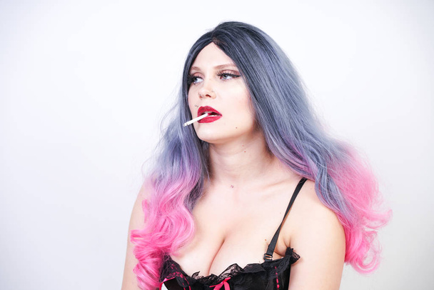 bad girl with piercing and long grey pink hair in black corset and torn fishnet pantyhose with mesh gloves sitting on the floor and Smoking cigarette on white background in Studio alone - Photo, image