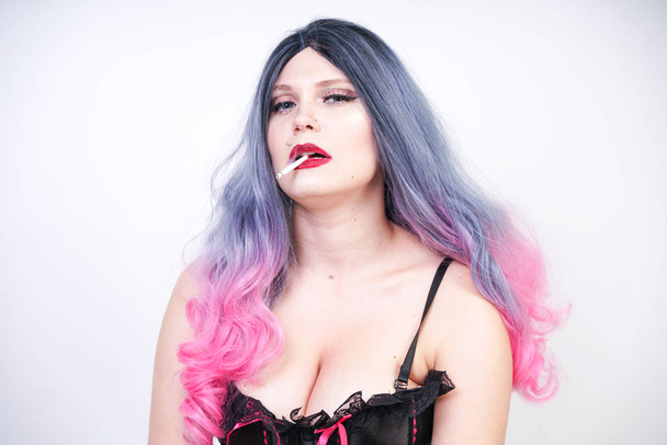bad girl with piercing and long grey pink hair in black corset and torn fishnet pantyhose with mesh gloves sitting on the floor and Smoking cigarette on white background in Studio alone - Fotó, kép