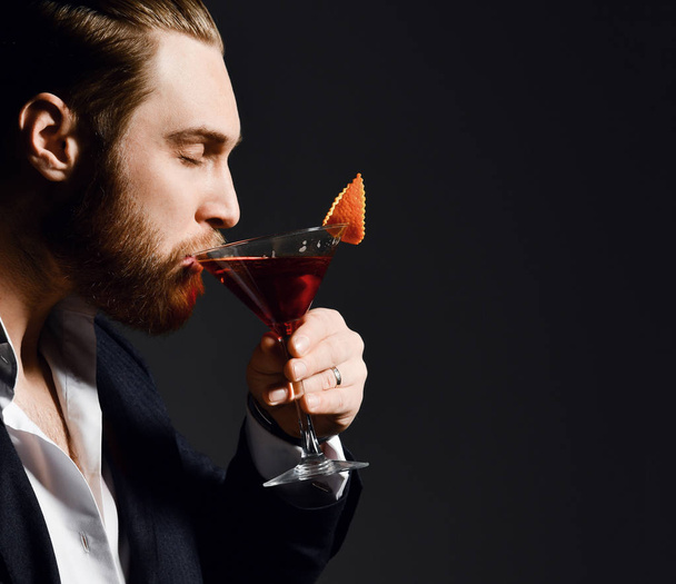 Brutal modern man in fashion suit bearded barman with closed in pleasure eyes drinks a delicious red cocktail with orange in martini glass - Zdjęcie, obraz